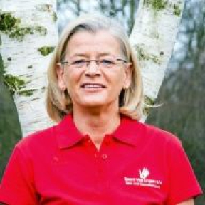 Eine blonde Frau mittleren Alters sitzt draußen vor einem hellen Baumhintergrund.